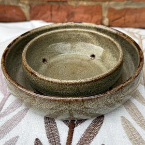 May include: A handmade, two-piece ceramic bowl set. The bowls are a speckled, earthy green with a darker brown rim. The smaller bowl has two small holes. The set is likely for serving or display.