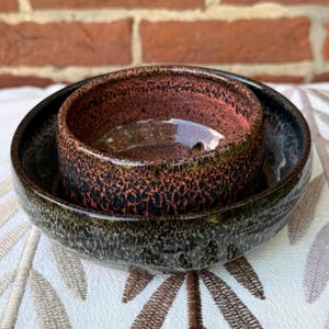 Puede incluir: Dos cuencos de cerámica, uno dentro del otro. El cuenco exterior es de color verde oscuro y negro, mientras que el cuenco interior es de color marrón rojizo con motas negras. Los cuencos están sobre una superficie blanca estampada.