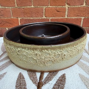 May include: A round ceramic bowl with a dark brown interior and a textured, light beige exterior featuring a porous, crater-like surface pattern. The bowl has a wide, shallow base and a smaller, elevated circular inner section in dark brown.