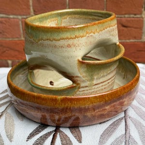 May include: A handmade ceramic yarn bowl featuring a wide, shallow base and a tall, cylindrical upper section with a deep, curved cutout for yarn. The pottery has a variegated glaze with earthy tones of sage green, cream, and deep rust-brown.