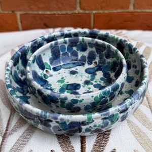 May include: Two nested ceramic bowls featuring a speckled pattern in shades of dark blue, light blue, and green on a white base. The bowls have a smooth, glossy finish with rounded edges and a circular shape, showcasing a hand-painted aesthetic.