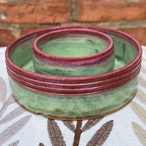 May include: A handmade ceramic serving bowl with a unique design. The bowl features a light green interior and exterior, with a vibrant red band around the rim. The bowl has a circular shape with a smaller, central bowl. The pottery is on a patterned surface.