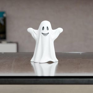 Peut inclure: Une figurine de fantôme blanc avec un visage souriant et les bras tendus. Le fantôme est posé sur une surface réfléchissante. L'arrière-plan est flou, avec une télévision et un mur de couleur neutre.