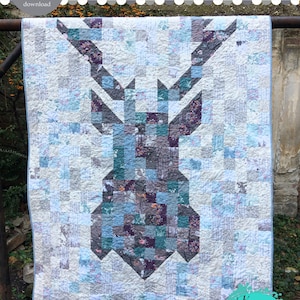 May include: A quilt pattern featuring a deer design in shades of blue, gray, and purple. The quilt is made of patchwork fabric squares and is displayed on a wooden frame. The text "DEAR DEER quilt pattern" is at the top of the image. The text "Katarina Roccella sewing patterns" is at the bottom of the image.
