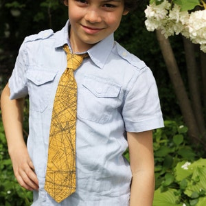 May include: A young boy wearing a light blue button-down shirt with a yellow and black patterned tie. He has curly brown hair and is smiling.