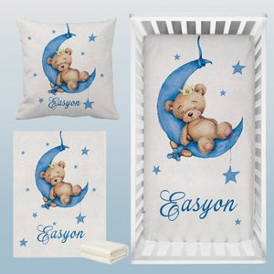 May include: A white crib bedding set with a blue crescent moon and a brown teddy bear wearing a gold crown. The set includes a fitted sheet, a pillowcase, and a blanket. The name "Easyon" is written in blue cursive on each piece.