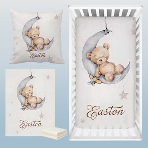 May include: A white crib sheet with a gray crescent moon and a brown teddy bear sleeping on it. The sheet has the name "Easton" written in brown cursive script. The crib sheet is part of a baby bedding set.