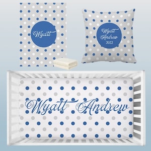 May include: A white crib with a blue and gray polka dot fitted sheet. The sheet has the name "Wyatt Andrew" written in a gray script font. There is a matching blue and gray polka dot pillow and blanket.