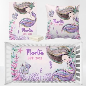 May include: A purple and pink crib bedding set with a mermaid theme. The bedding features a mermaid with long brown hair, a purple tail, and a crown. The bedding also includes a pillow with the name "Martin" and the year "EST. 2021" printed on it.