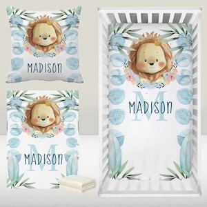 May include: A blue and white crib bedding set with a lion design. The set includes a fitted sheet, a crib blanket, and a pillow. The bedding features a watercolor-style illustration of a lion with a crown, surrounded by blue leaves and flowers. The name "MADISON" is written in blue letters on the bedding.