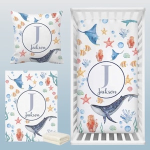 May include: A white crib bedding set with a blue whale, starfish, and coral reef design. The set includes a fitted sheet, a pillowcase, and a blanket. The bedding is personalized with the letter "J" and the name "Jackson".