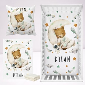 May include: A white crib bedding set with a moon and stars theme. The set includes a fitted sheet, a crib blanket, and a pillow. The bedding features a brown bear sleeping on a crescent moon with a green and orange rocket and stars. The bedding is personalized with the name "DYLAN".