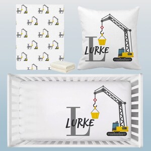 May include: A white crib sheet with a yellow construction crane design. The crane is lifting a yellow bucket and has the name "Lurke" written in gray letters.