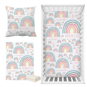 May include: A set of baby bedding featuring a rainbow pattern with pink, yellow, and blue colors. The set includes a crib sheet, a blanket, and a pillow. The name "Annabelle" is written on each piece of bedding.