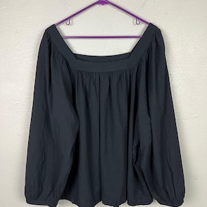 May include: A black long-sleeve top with a square neckline, hanging on a purple hanger. The top has a gathered detail below the neckline and a loose, flowing fit. The sleeves are long and end in a gathered cuff.
