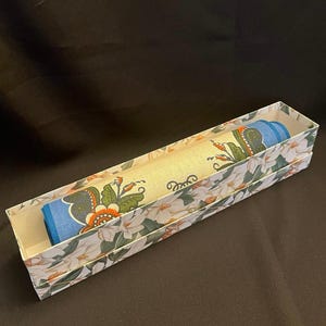May include: A rolled-up, patterned textile is presented inside a rectangular box. The textile features a floral design with orange, green, and blue accents. The box is decorated with a white floral pattern on a light background.