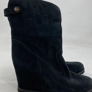 May include: A black suede wedge boot featuring a mid-calf height, a rounded toe, and a thick rubber sole. The upper shaft includes a decorative strap secured by a circular metallic button embossed with the UGG logo, providing a distinct profile.