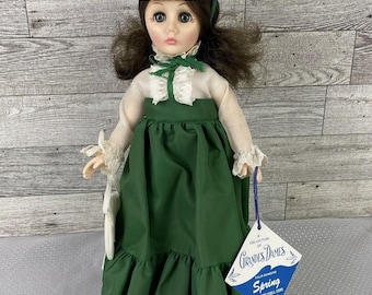 May include: A vintage doll wearing a green dress with white lace trim and a straw hat adorned with a green bow and yellow flowers. The doll has brown hair and is holding a small white purse. A blue tag reads "Grandes Dames Spring".