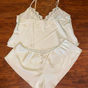 May include: A light mint green two-piece pajama set consisting of a camisole and shorts. The camisole features thin adjustable shoulder straps and a V-neckline trimmed with white floral lace. The shorts have an elasticized waistband.