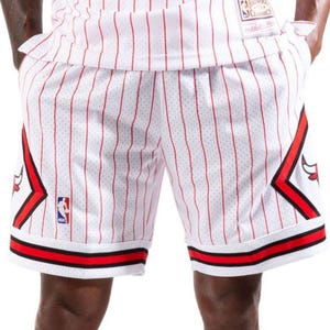 May include: White basketball shorts featuring thin vertical red pinstripes, a red and black diamond-shaped graphic on each side with a bull logo, a small NBA logo patch on the right leg, and thick red and black horizontal stripes at the hem.