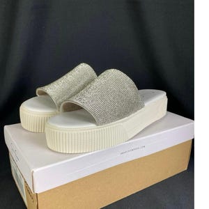 May include: Silver rhinestone slide sandals with a white platform sole. The sandals are on a white box with the brand name Jessica Simpson. The platform sole has a ridged texture. The background is black.