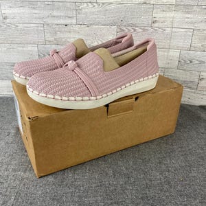 May include: A pair of slip-on shoes in a light pink hue, featuring a woven texture and a white sole with decorative stitching. The shoes are displayed on a brown cardboard box, suggesting a new product.