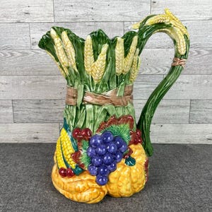 May include: A ceramic pitcher with a harvest theme. The pitcher is decorated with corn, grapes, and pumpkins in shades of yellow, green, and purple. The handle is green and shaped like wheat.
