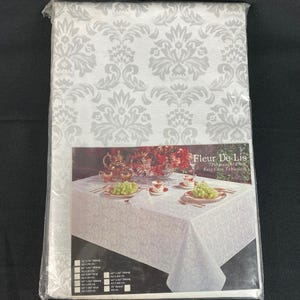 May include: A white tablecloth with a silver damask pattern. The tablecloth is packaged in plastic and labeled "Fleur De Lis Perma-Press Easy Care Tablecloth".