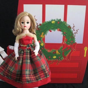 May include: A vintage doll in a red and green plaid dress with a red bodice and white gloves. The doll has blonde hair and is standing in front of a red door decorated with a Christmas wreath.