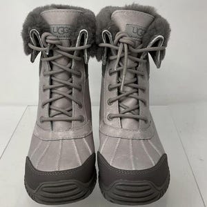 May include: A pair of grey lace-up boots featuring a dark grey rubber toe cap, a grey leather upper, and a plush grey shearling cuff at the collar. The tongue of each boot displays the UGG Australia logo patch with matching grey laces.
