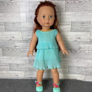 May include: A doll with auburn hair and blue eyes wearing a turquoise pleated dress and matching shoes with pink bows. The doll has a yellow collar and is standing in front of a gray and white wooden wall.