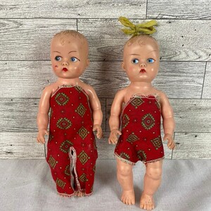 May include: Two vintage baby dolls with painted features and blue eyes. They wear red patterned rompers with a white trim. One doll has a yellow bow in its hair. The dolls are standing on a white surface.
