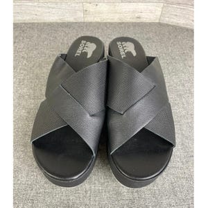 May include: Pair of black Sorel platform sandals with crisscross straps. The sandals have a textured design on the straps and a white sole. The Sorel logo is visible on the footbed.