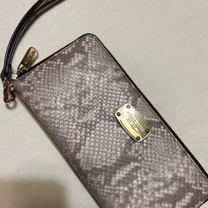 Può includere: Un portafoglio rettangolare con cinturino da polso caratterizzato da un motivo a pelle di serpente grigio e bianco. Include una zip in metallo dorato e un cinturino abbinato. Una placca rettangolare dorata reca l'incisione Michael Kors.