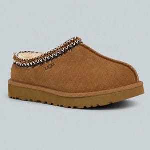 May include: A tan suede slip-on slipper with a thick brown rubber sole and a cream-colored shearling lining. The collar features a decorative brown and white woven trim, and the UGG brand logo is embossed on the side of the upper exterior.
