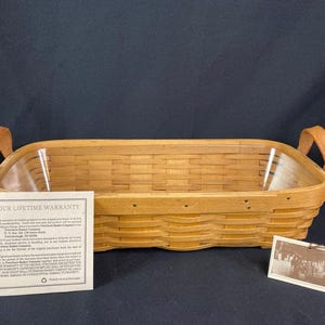 May include: A rectangular woven wood basket with two tan leather handles and a clear plastic liner. A rectangular paper card titled YOUR LIFETIME WARRANTY and a small rectangular tag with a sepia-toned photograph are placed in front of the basket.
