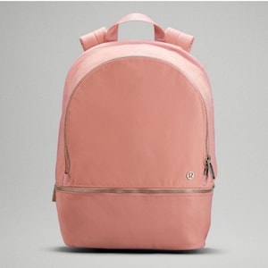 May include: A dusty pink backpack featuring a large main compartment with a curved zipper closure, a smaller horizontal zippered pocket at the base, a top handle, and adjustable shoulder straps. A small circular logo is visible on the front.