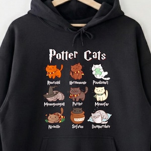 May include: Black hooded sweatshirt with a front graphic that reads "Potter Cats" in a stylized font. The graphic features cartoon cat illustrations with names like "Ronweasley" and "Dumbledore". The sweatshirt has a front pocket and a drawstring hood.