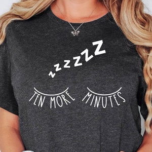 May include: Dark gray t-shirt with white text and graphic. The shirt features the words "Ten More Minutes" below a pair of closed eyelashes and the sound "zzzzz" above. The shirt is a casual, comfortable style.