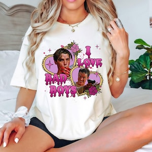 May include: Cream-colored t-shirt featuring a graphic design with the text "I LOVE BAD BOYS" in pink, with two images of men inside heart-shaped frames. The design includes pink roses and stars.