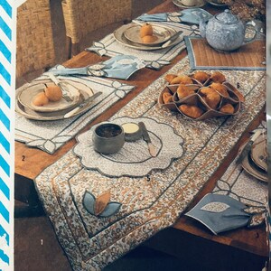 May include: A Simplicity sewing pattern for a table runner and placemats. The pattern is number 6229 and is designed by Tricia Guild. The pattern features a floral design in shades of brown, beige, and blue. The pattern is shown on a wooden table with a teapot, cups, and saucers.