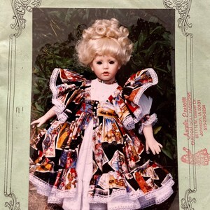 May include: A doll wearing a colorful dress with white lace trim. The doll has blonde hair and blue eyes. The image is from a pattern for a doll dress called "The "Love" Patterns" by Angela Coriatti. The pattern is for a 28 inch doll and costs $7.95.