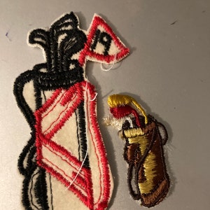May include: Two embroidered felt patches depicting golf bags. The larger patch is white with red and black stitching, and the smaller patch is brown with gold stitching.