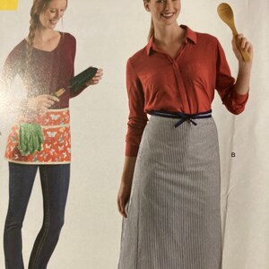 May include: Two women wearing different aprons. The woman on the left is wearing a red long-sleeved shirt and a floral apron with pockets. The woman on the right is wearing a red long-sleeved shirt and a black and white striped apron with a blue ribbon.