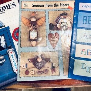 May include: Three craft books with colorful covers. The first book is titled "Farm Welcomes" and features a blue door with a welcome sign. The second book is titled "Seasons from the Heart" and features a heart-shaped design with an angel. The third book is titled "Alphabets and Monograms" and features a blue background with the letter "A" in white.