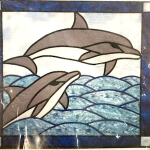 Puede incluir: Un panel de vidriera con dos delfines saltando sobre olas azules. Los delfines son grises con detalles blancos, sobre un cielo nublado. El panel está enmarcado con un borde azul oscuro.