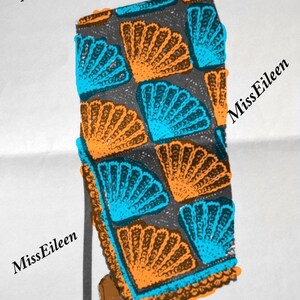 May include: A crocheted throw blanket with a fan-shaped pattern in shades of orange, blue, and gray. The blanket is leaning against a brown armchair with a checkered throw draped over it. The text "Miss Eileen's Vintage" and "Miss Eileen" is visible in the image.