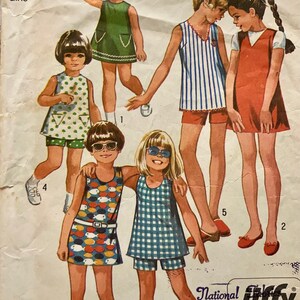 May include: A vintage sewing pattern for girls' dresses and shorts. The pattern includes five different styles, with options for a green dress with a white trim, a striped dress with red shorts, a red dress with a white trim, a dress with a fish print, and a gingham dress with blue shorts. The pattern is from Simplicity and is number 3865.