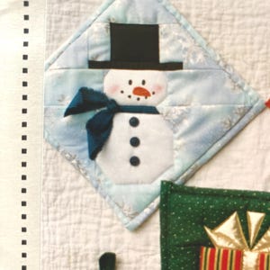 May include: A quilted Christmas-themed potholder featuring a snowman design. The snowman has a black top hat, a blue scarf, and a carrot nose. The background is light blue with snowflake patterns. The potholder is diamond-shaped.