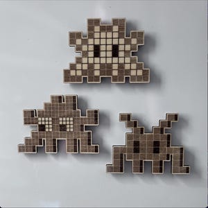 Op de afbeelding: Drie houten pixel art space invaders in bruin en wit. De space invaders staan in verschillende poses, een kijkt naar voren, een naar links en een naar rechts.